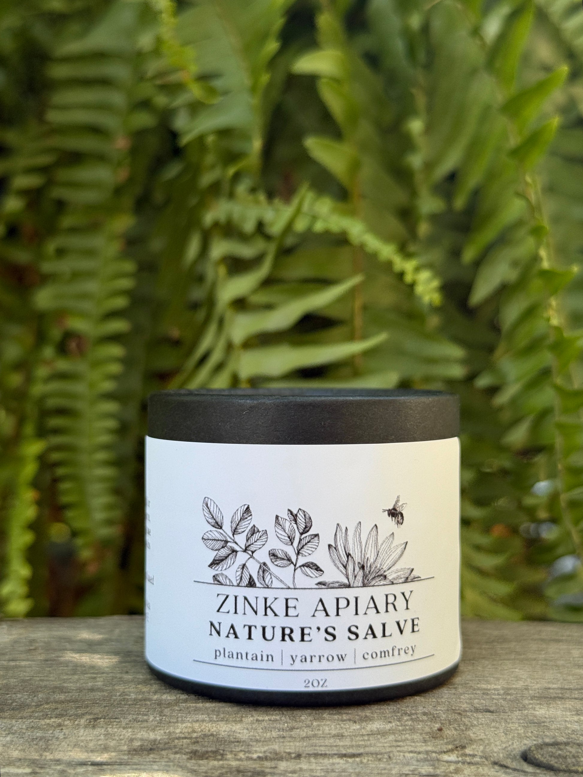 Zinke Apiary Nature's Salve jar with a black lid on a wooden surface with green foliage in the background